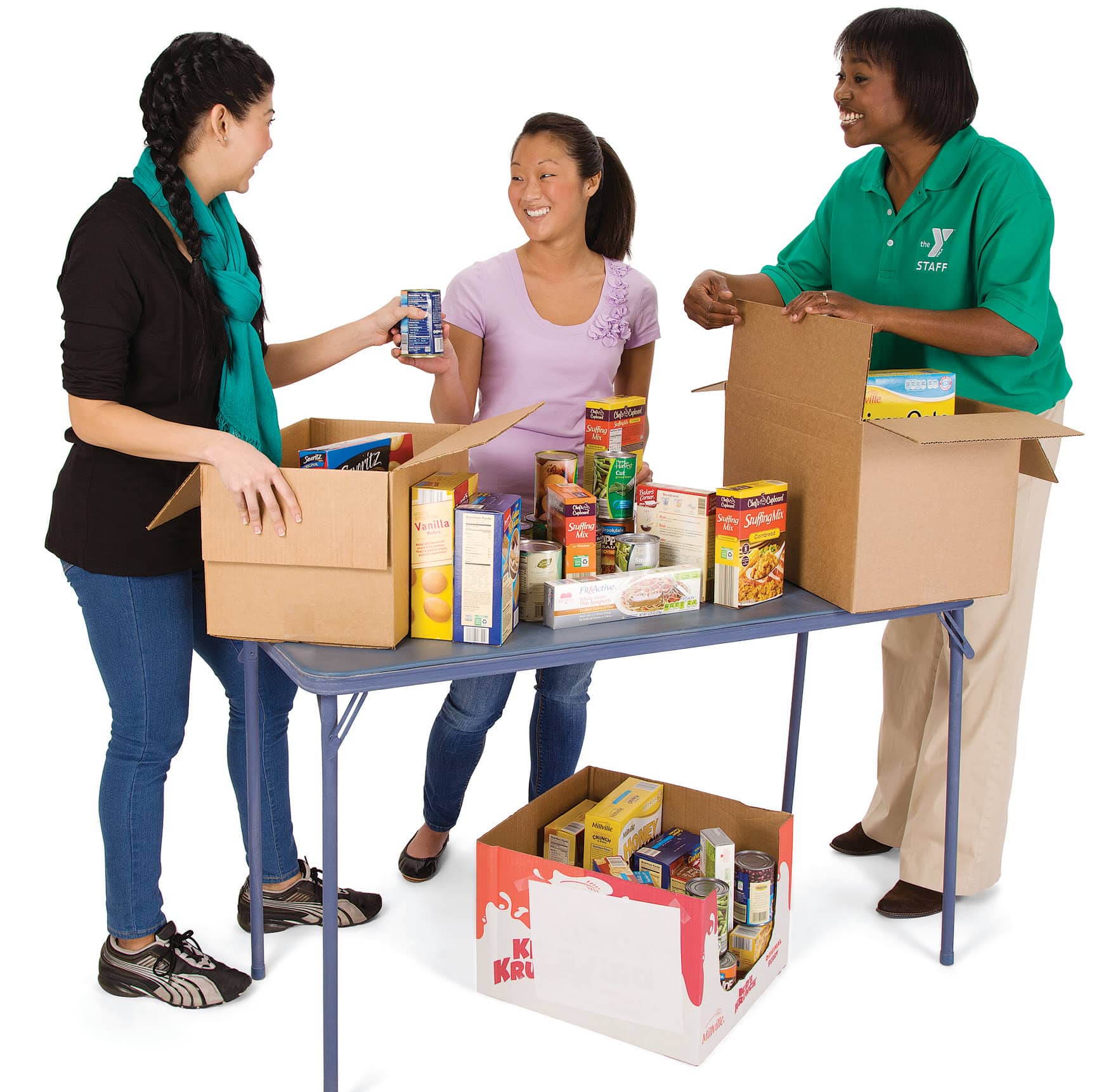 The image depicts three women participating in a food drive organization activity. They are standing around a folding table that holds several open cardboard boxes with various non-per