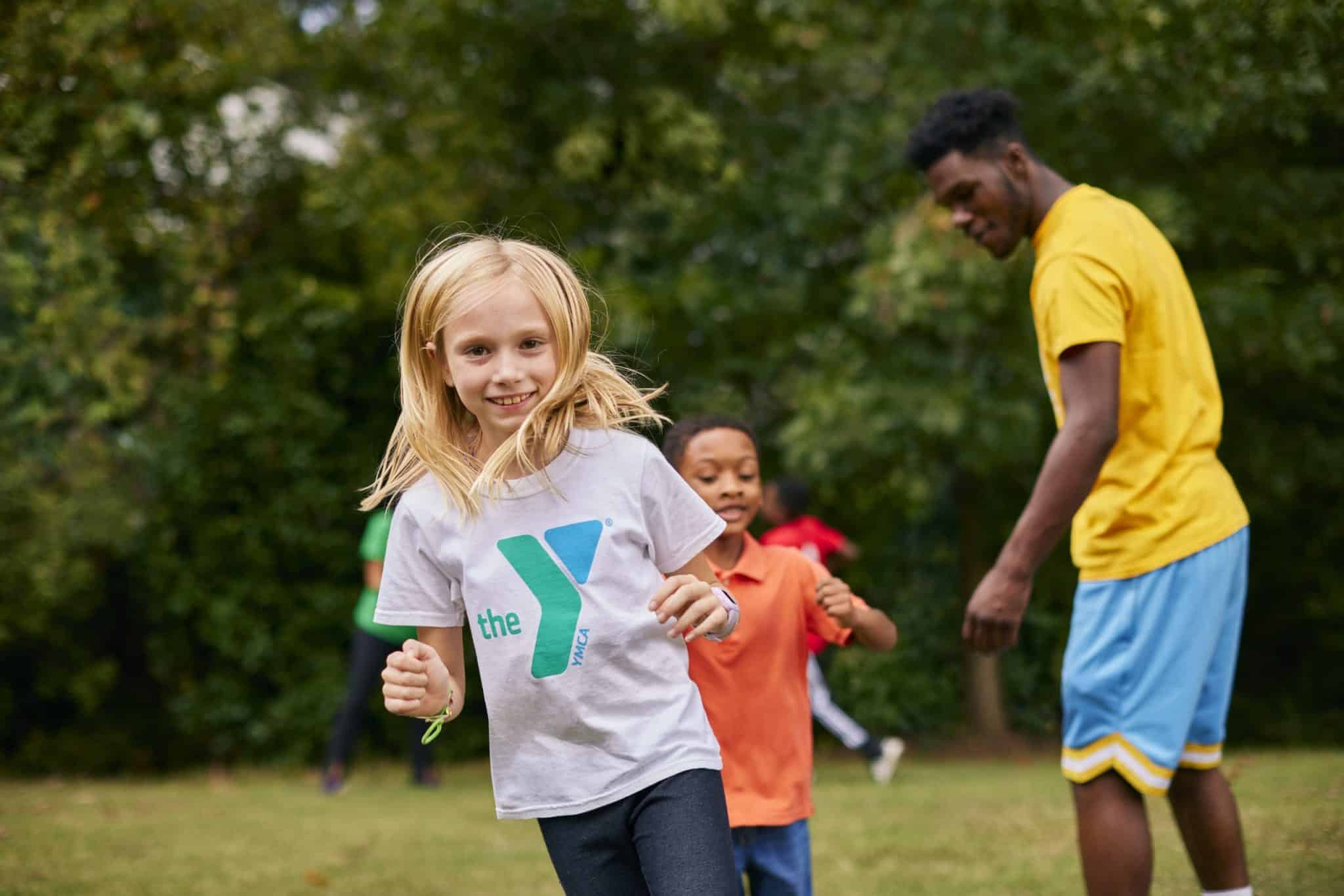 This image captures a cheerful, active scene in an outdoor setting with greenery. A young girl with blonde hair wearing a gray T-shirt featuring the YM