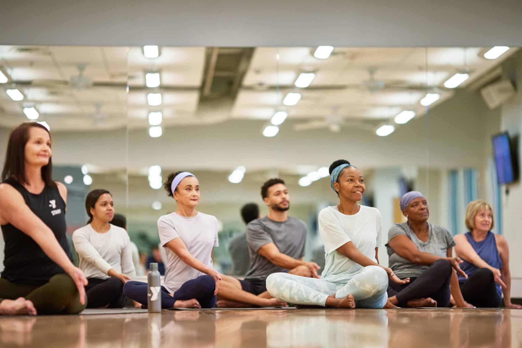 The image shows a group of seven adults in a yoga or fitness studio, participating in what appears to be a meditation or relaxation class. They are seated