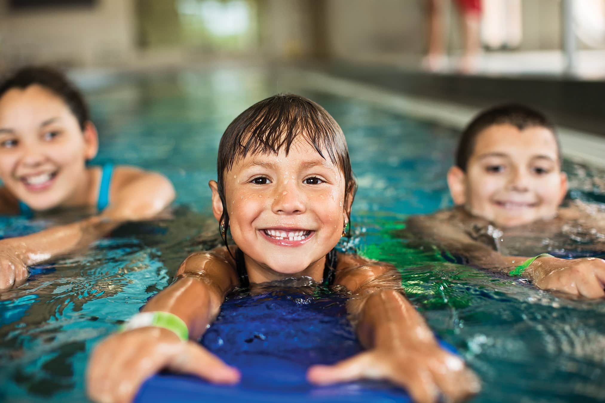 The image captures three children having fun in an indoor swimming pool. The child in the center is looking directly at the camera with a joyful expression, showcasing