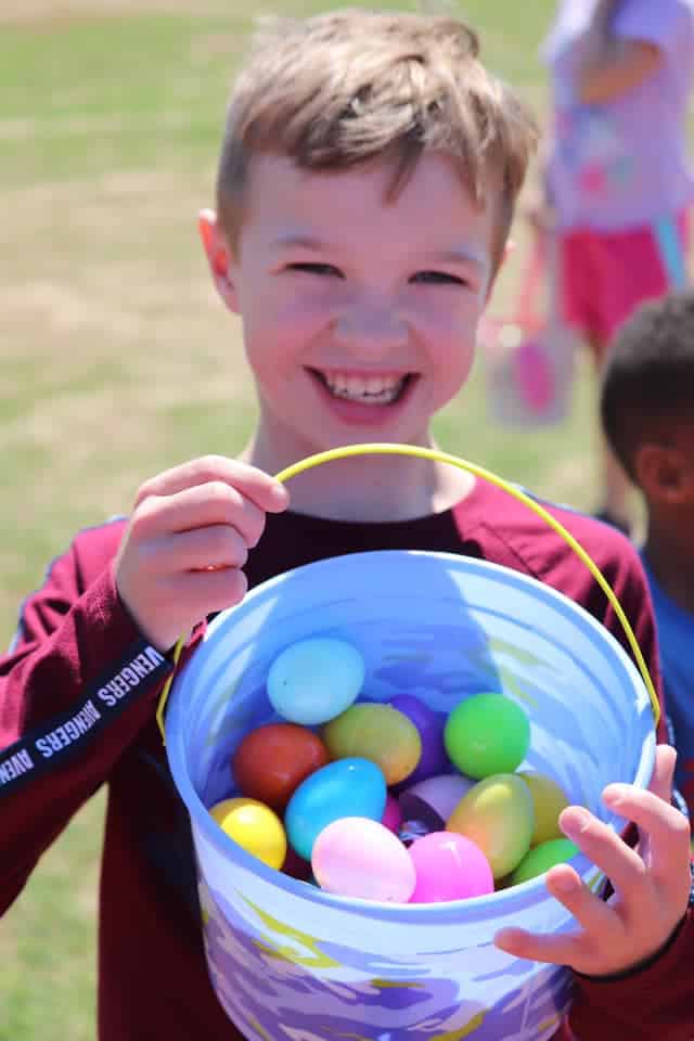 This image features a young boy outdoors on a sunny day, holding a large bucket filled with colorful plastic eggs. He has a joyful expression on his face