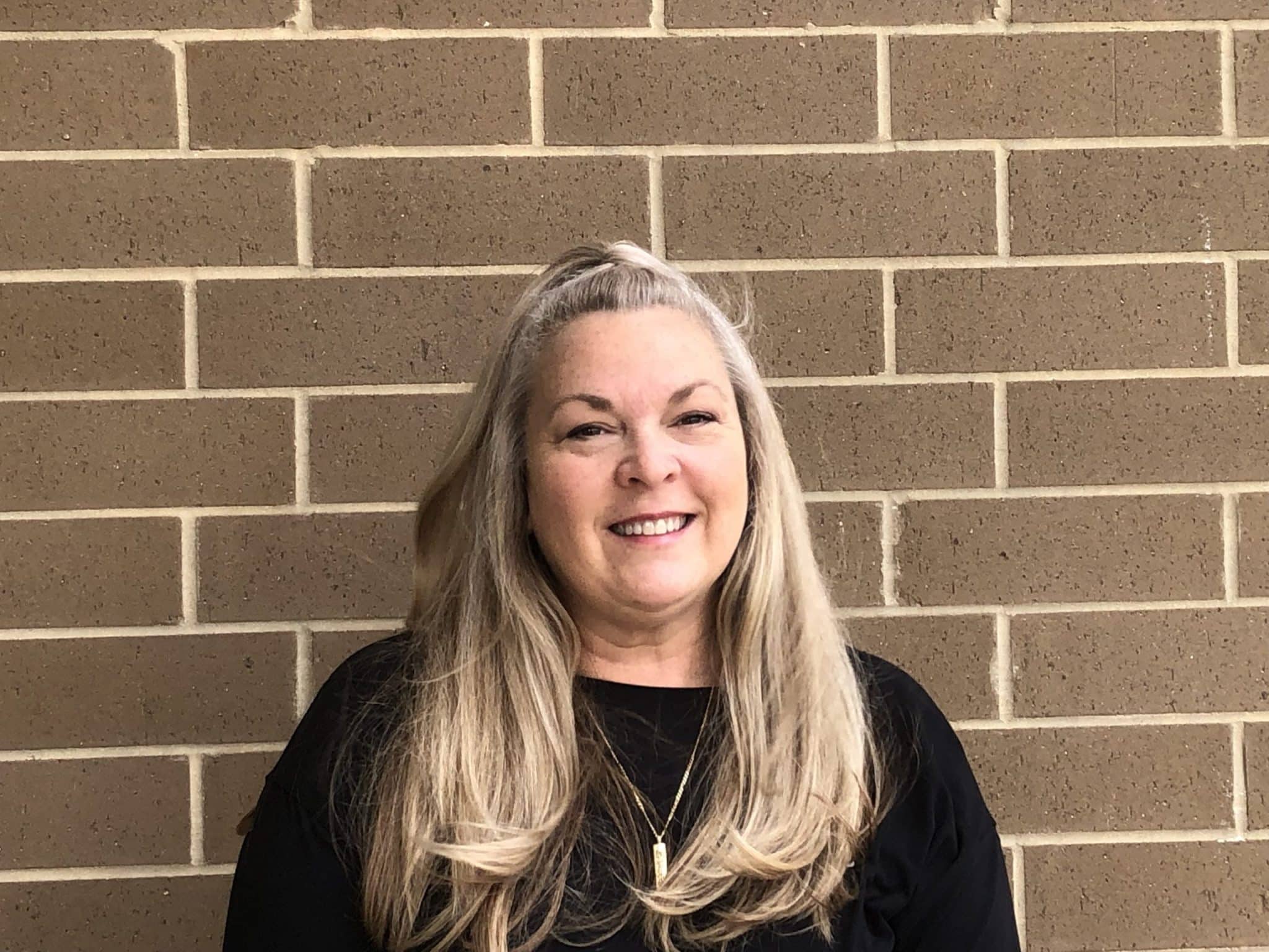 This image features a woman smiling at the camera. She has long, blonde-gray hair and is wearing a black top. The background is a reddish