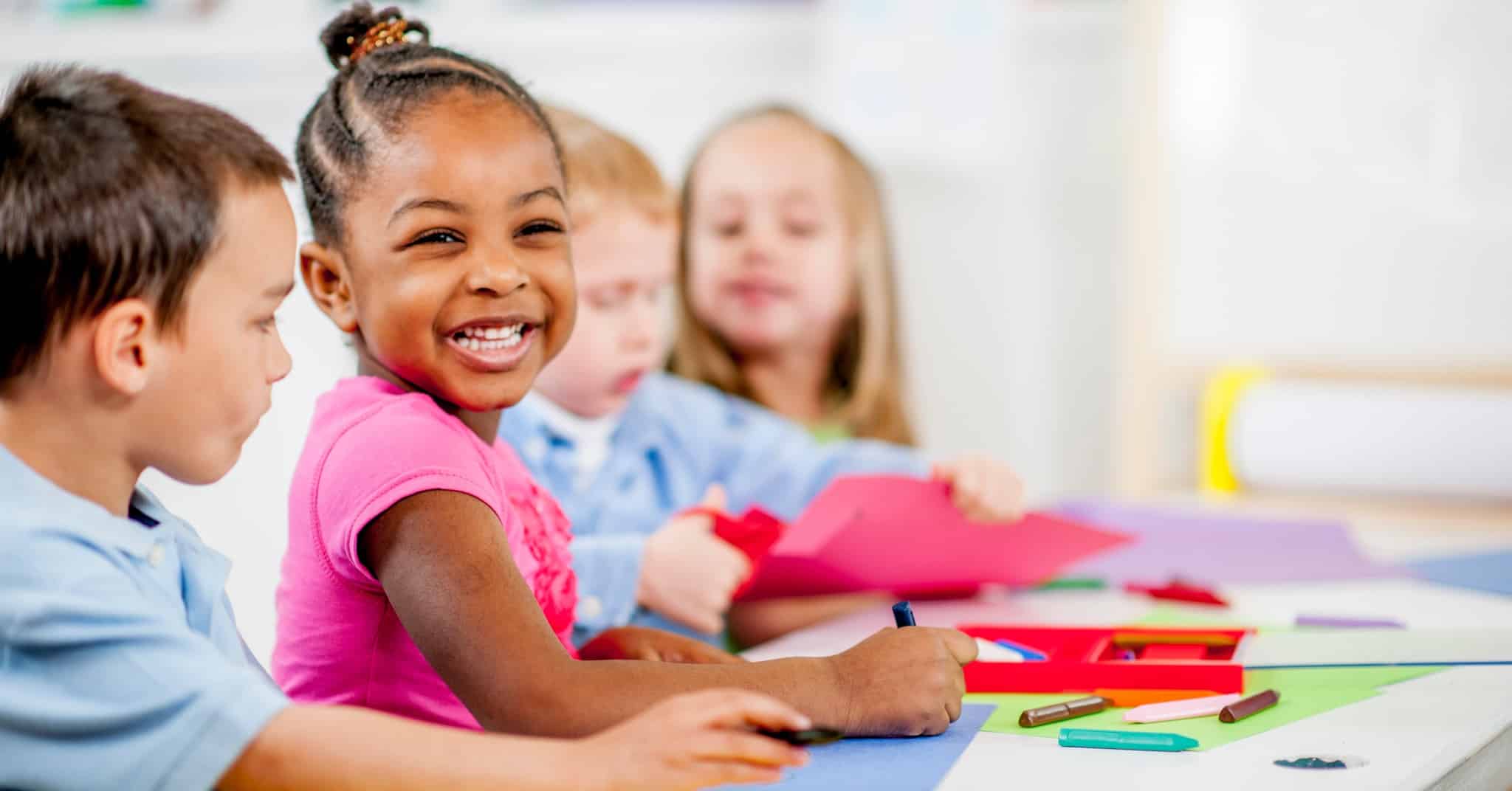 This image features a cheerful scene with a group of young children engaged in a classroom activity. A young girl with a bright smile, wearing a pink top