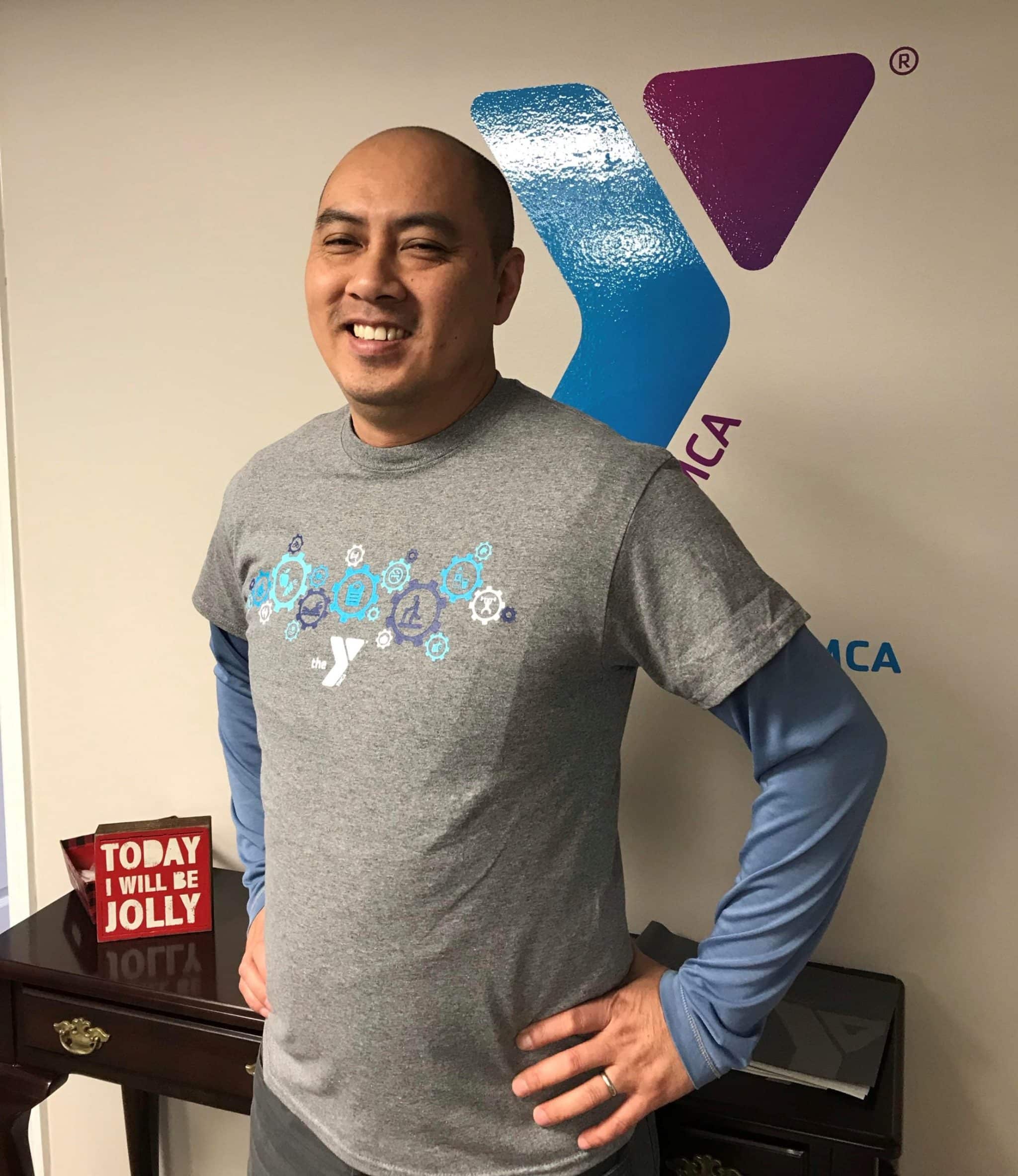The image features a man standing indoors against a plain white wall that has a colorful logo with the letter "Y" and some triangles in blue and purple