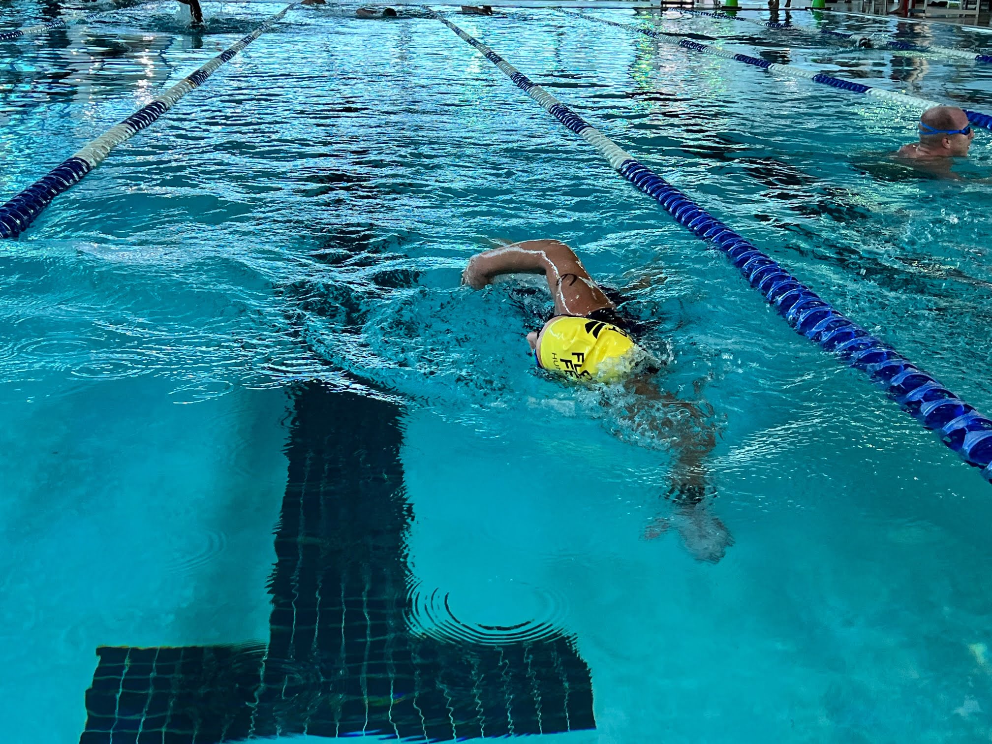 The image shows an indoor swimming pool where two swimmers are actively engaged in swimming laps. One swimmer, wearing a yellow swim cap marked with the