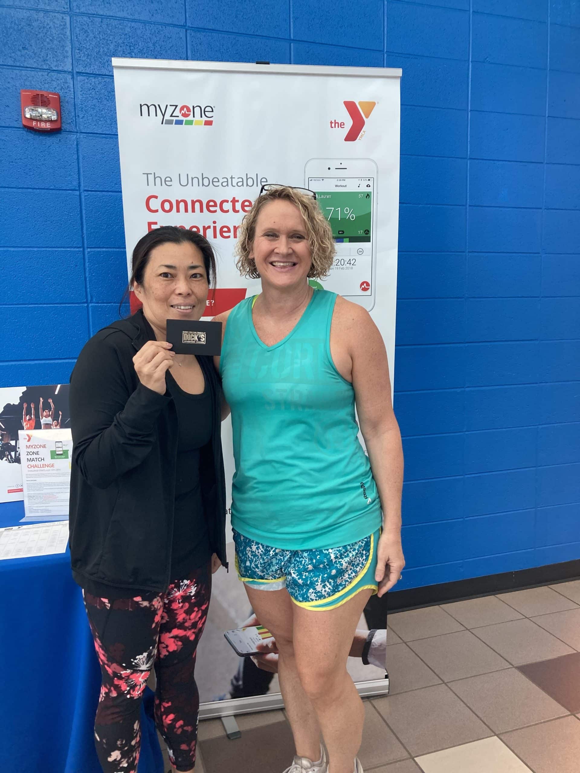 The image features two smiling women posing in front of a promotional banner for Myzone at a YMCA facility, as indicated by the logos and information on