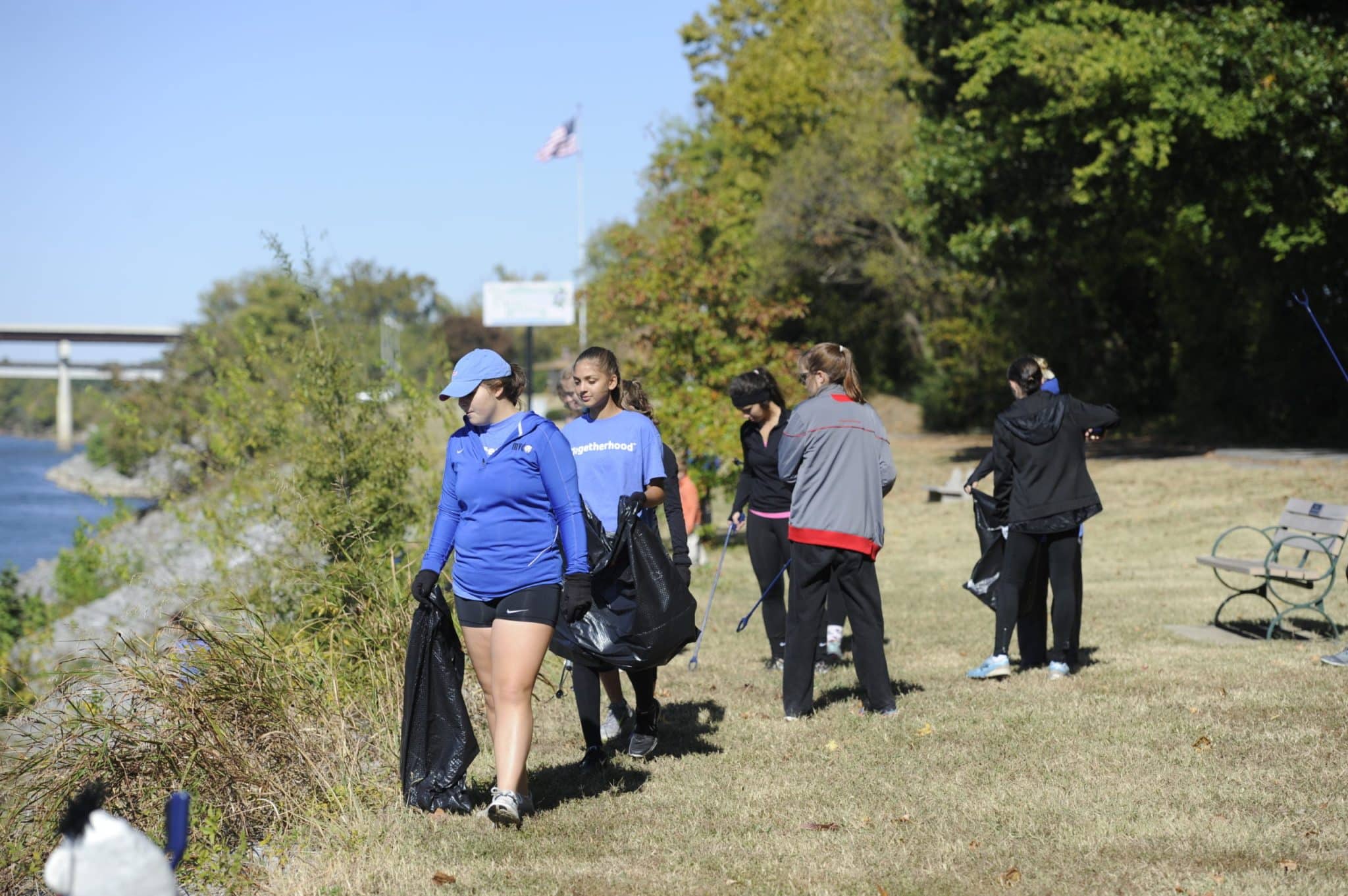 This image shows a group of people involved in a community clean-up activity near a riverbank. The individuals, wearing casual and athletic attire along with gloves