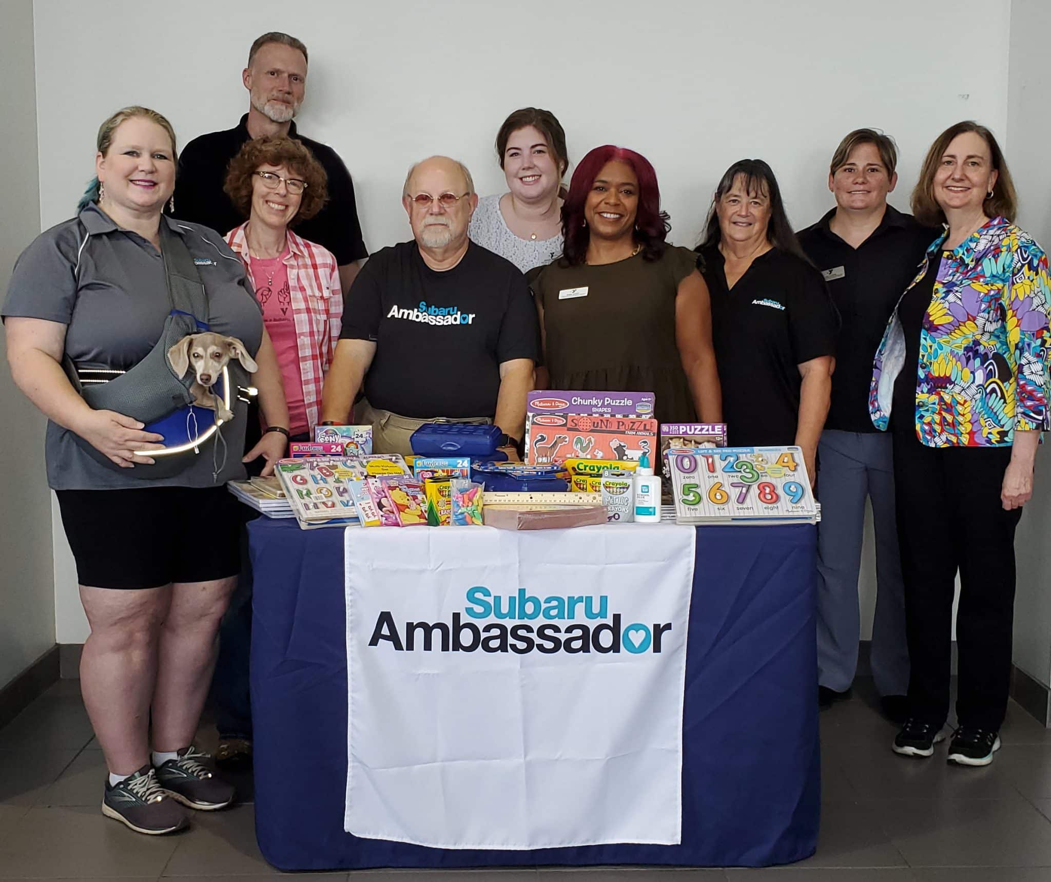 The image features a group of eight people, consisting of four women and four men, posing behind a table draped with a "Subaru Ambassador" table