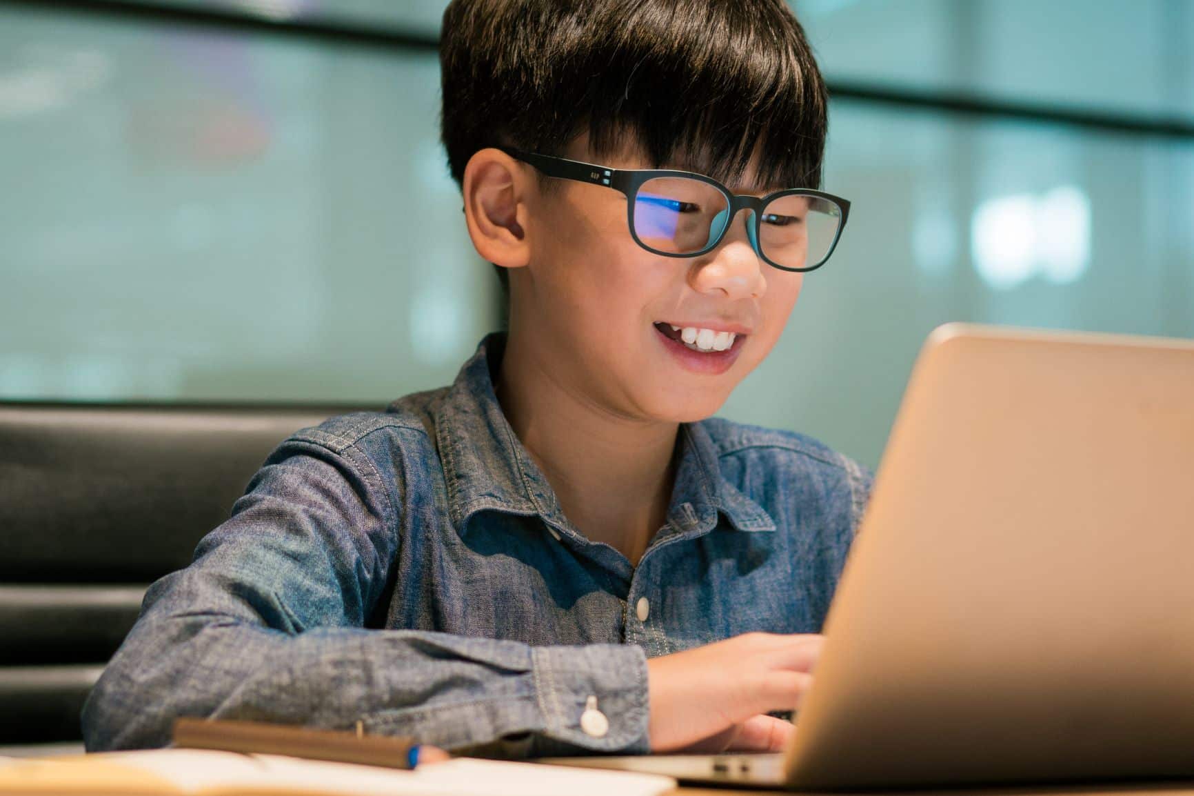 This image displays a young boy wearing glasses and a denim shirt, smiling joyfully as he looks at a laptop screen. The setting appears to be a