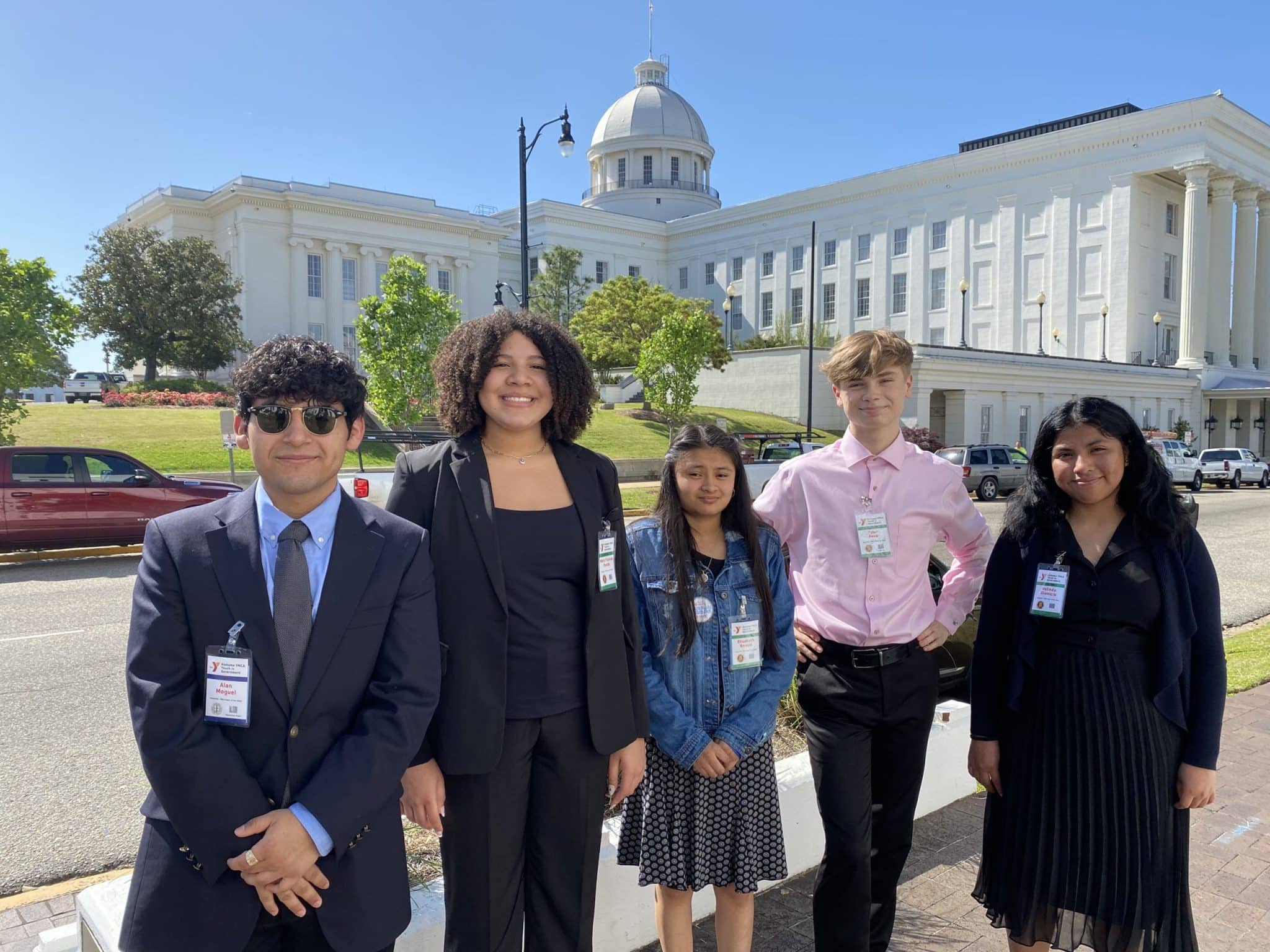 This image features five young individuals posing in front of a large, white building with classical architectural features including a central dome and tall columns. The setting appears