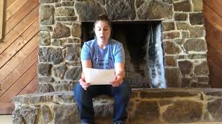 This image shows a woman sitting in front of a large stone fireplace. She is holding a piece of paper and appears to be reading from it. The