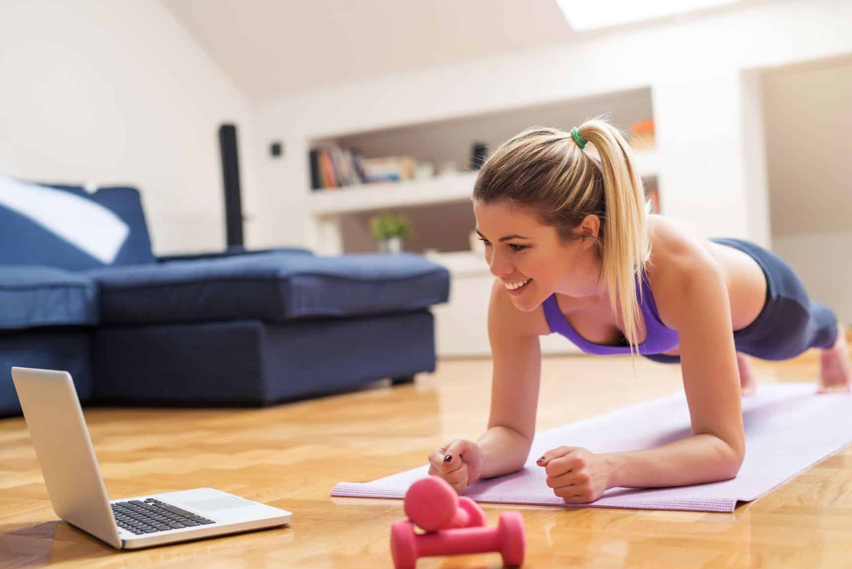 The image shows a young woman exercising in a home setting. She is in a plank position on a pink mat, smiling and looking at a laptop that