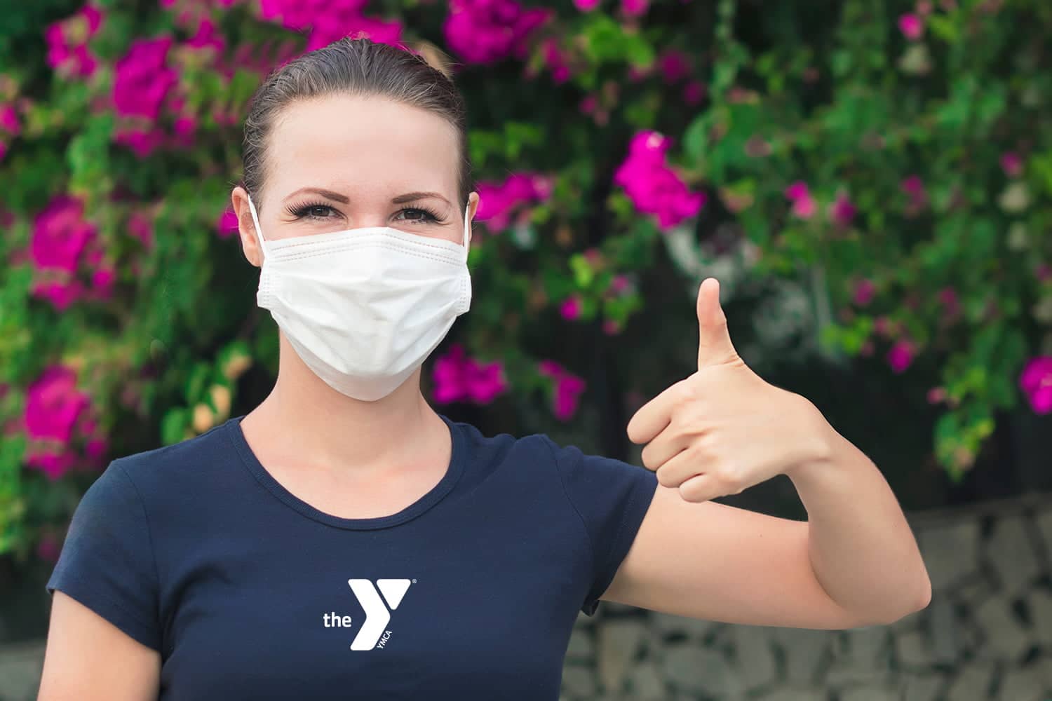 This image features a woman outdoors wearing a white face mask. She is giving a thumbs-up sign with her right hand, which can symbolize approval or