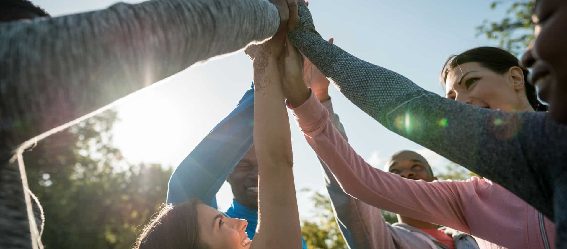 This image features a diverse group of adults outdoors, participating in a team-building activity. They are all smiling and have their hands joined together above their heads