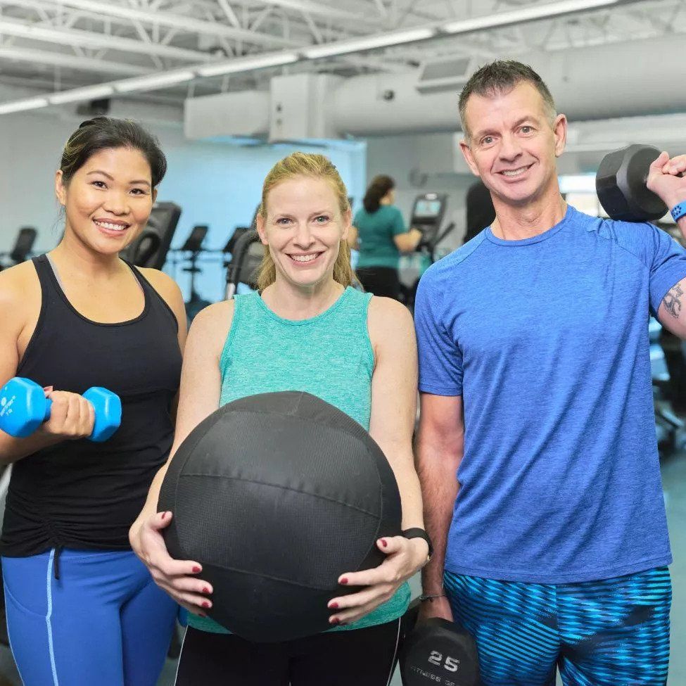 The image features three adults in a gym setting, each holding different pieces of exercise equipment and smiling at the camera. On the left, a woman in