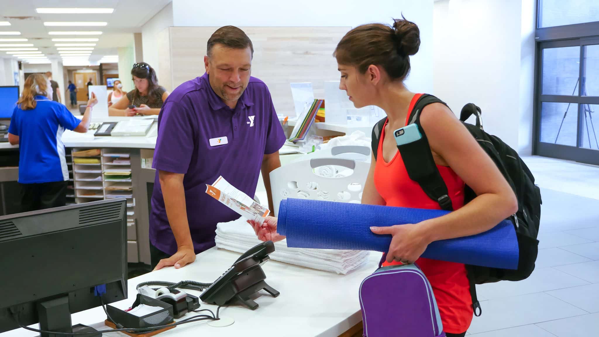 This image depicts a customer service interaction at a gym or fitness center. A man dressed in a purple polo shirt embellished with a "Y" logo