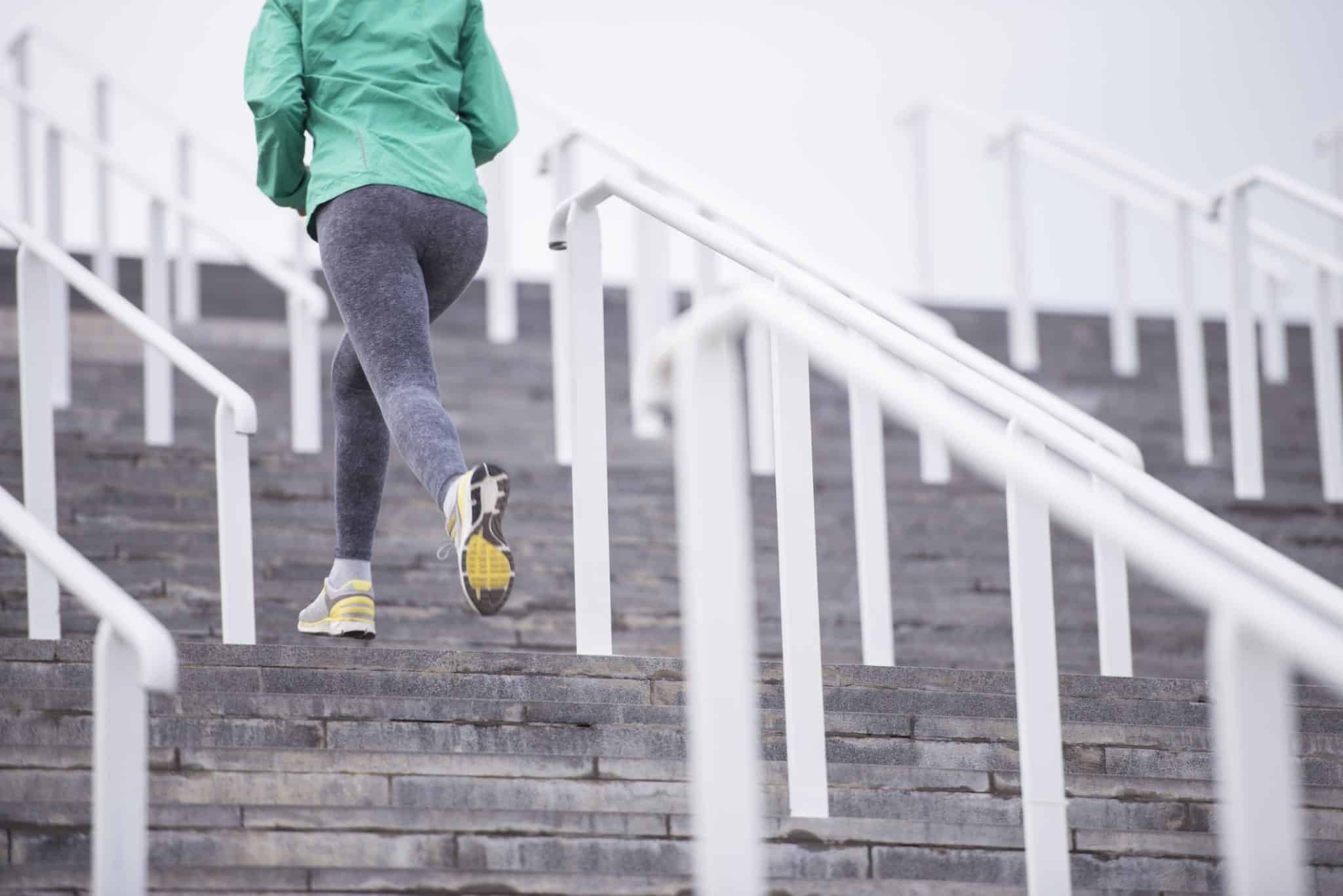 Unrecognized athlete woman running up on stairs, wearing running shoes, green sport rain jacket and grey leggings. Fitness, diet, active lifestyle concept.